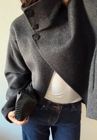 Manteau enveloppant gris en mélange de laine avec un col montant et trois boutons noirs. Associé à un haut blanc et un pantalon en denim foncé. Sac à main noir texturé en main.