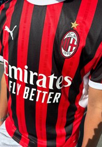 Camisa de futebol do AC Milan com listras verticais vermelhas e pretas, logótipo branco "Emirates", colarinho preto e emblema do clube com uma estrela dourada.