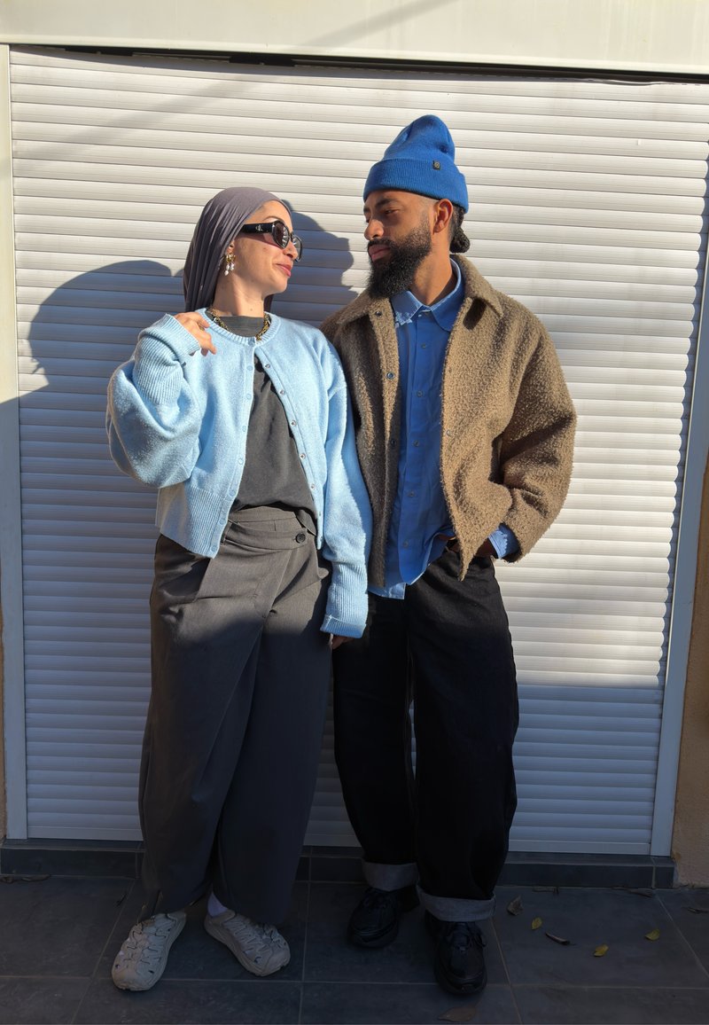 Két ember áll közel egymáshoz, egymásra néznek; az egyik világoskék kardigánt és fejkendőt visel, a másik kék sapkát és barna kabátot.