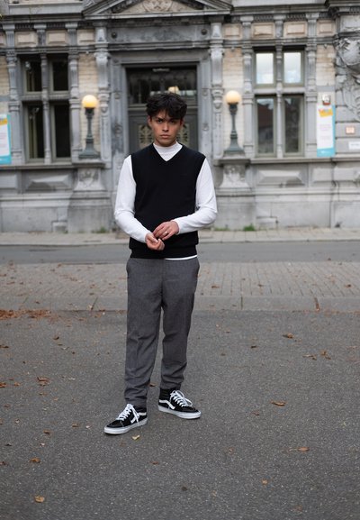 Suéter negro sin mangas sobre una camisa de manga larga blanca, acompañado de pantalones grises y zapatillas Vans negras. Entorno urbano al aire libre.