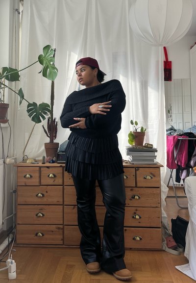Suéter negro de canalé con diseño de hombros al descubierto, con un fondo de capas plisadas; combinado con pantalones acampanados negros brillantes y zapatos de slip-on color bronce. Ambiente interior.