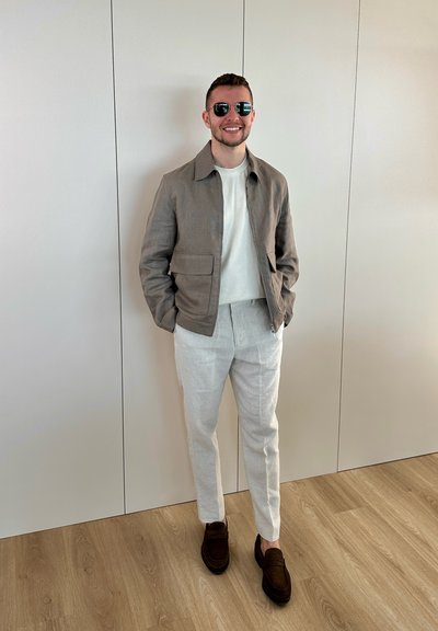 Hombre sonriendo, con gafas de sol, chaqueta marrón claro, camisa blanca, pantalones beige claros y mocasines marrón oscuro, de pie sobre suelo de madera junto a una pared lisa.