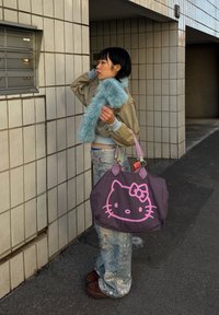Jeune femme portant une veste courte, une écharpe et un jean large, tenant un sac Hello Kitty violet, debout près d'unités de boîtes aux lettres carrelées.