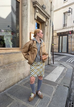 Chaqueta marrón sobre una sudadera con capucha gris, minifalda estampada, calcetas grises hasta la rodilla y zapatillas multicolores. Los accesorios incluyen gafas de sol y un bolso pequeño.