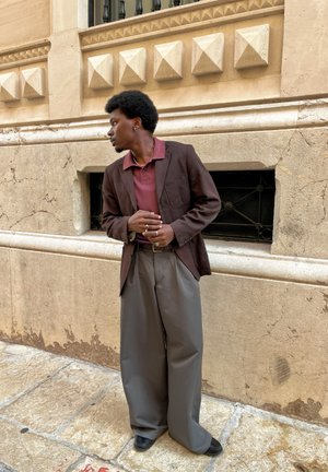 Blazer marrón sobre camiseta polo granate, combinado con pantalones grises sueltos y zapatos negros. Fondo arquitectónico con pared texturizada.