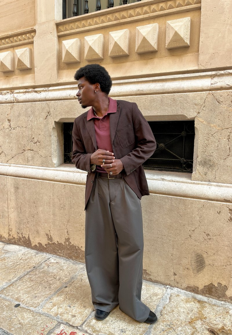 Blazer marrone sopra una polo bordeaux, abbinato a pantaloni grigi larghi e scarpe nere. Sfondo architettonico con muro testurizzato.