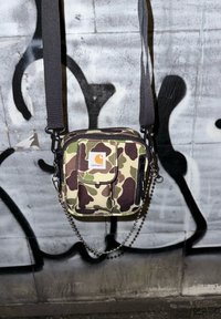 Camouflage crossbody bag with black zip, adjustable strap, and chain detail. Front flap pocket and fabric texture.