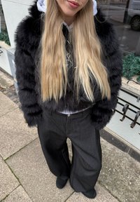 Jachetă neagră din blană sintetică cu design scurt, asortată cu pantaloni gri cu picioare evazate și un hanorac alb dedesubt. Texturi netede și culori solide.