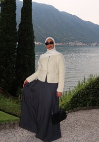 Chaqueta blanca con cremallera y cinturón, combinada con una falda plisada gris. Los accesorios incluyen gafas de sol negras y un pequeño bolso negro.