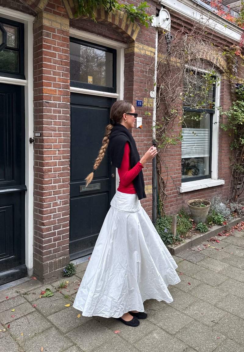 Lange witte rok met een uitlopende vorm, gecombineerd met een rood langemouwen top en een zwarte sjaal. Haar in een vlecht. Bakstenen muur en planten op de achtergrond.