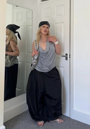 Mujer con pañuelo negro en la cabeza, top gris sin mangas con drapeado, bolso, pantalones anchos negros, pulsera plateada y sandalias, de pie cerca de una puerta blanca y un espejo.