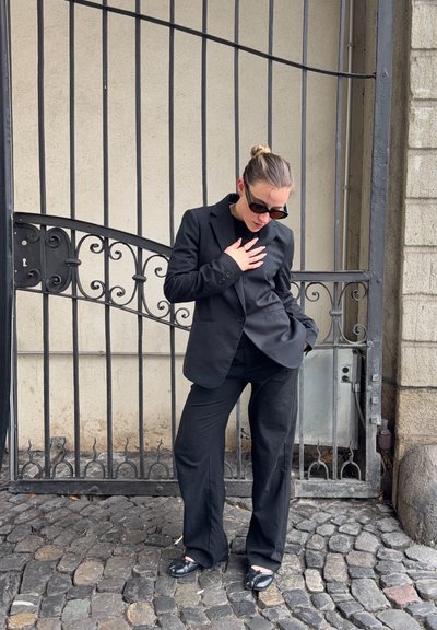 Mujer con traje negro y gafas de sol está de pie en una calle empedrada, tocándose el pecho con una mano, junto a una puerta de metal ornamentada.