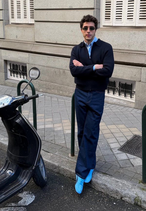 Νεαρός άνδρας με γυαλιά ηλίου, μαύρο μπουφάν, μπλε παντελόνι και φωτεινά μπλε παπούτσια, στηριγμένος σε ένα κράσπεδο κοντά σε ένα παρκαρισμένο μαύρο σκούτερ.