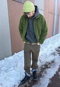 Homme portant un bonnet vert et une veste, un sweat à capuche noir, un pantalon marron et des bottes noires, debout sur un trottoir enneigé devant un mur multicolore.