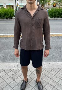 Braun gestreiftes Langarmhemd mit einer Brusttasche, kombiniert mit marineblauen Shorts. Schwarze Schuhe runden das Outfit ab. Die Textur wirkt leicht.
