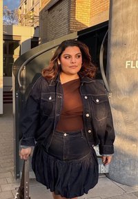Mujer con chaqueta de mezclilla negra y falda, con una blusa marrón, caminando afuera cerca de la entrada de un edificio mientras sostiene un bolso.
