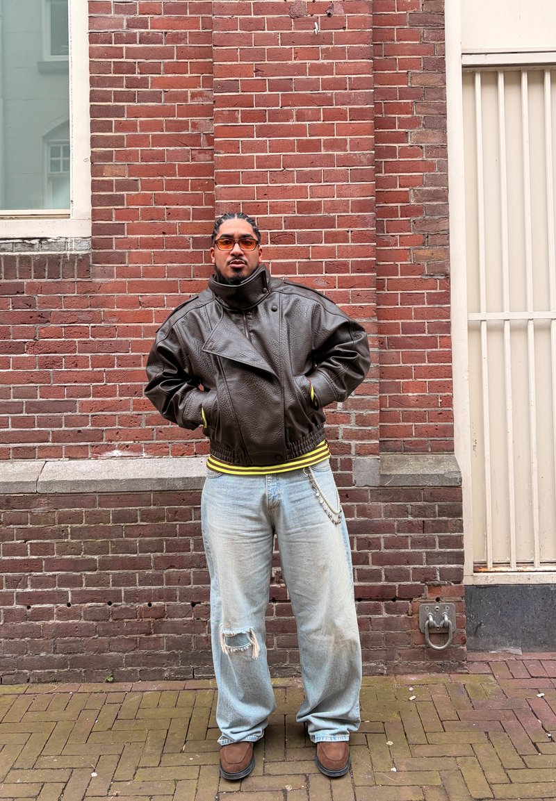 Homme aux cheveux tressés portant des lunettes teintées orange, une veste en cuir marron, un jean clair déchiré et des chaussures marron, debout contre un mur en briques rouges.