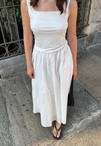 Top bianco senza maniche con scollatura quadrata, abbinato a una gonna midi bianca fluida con dettagli a laccio. Infradito nere.
