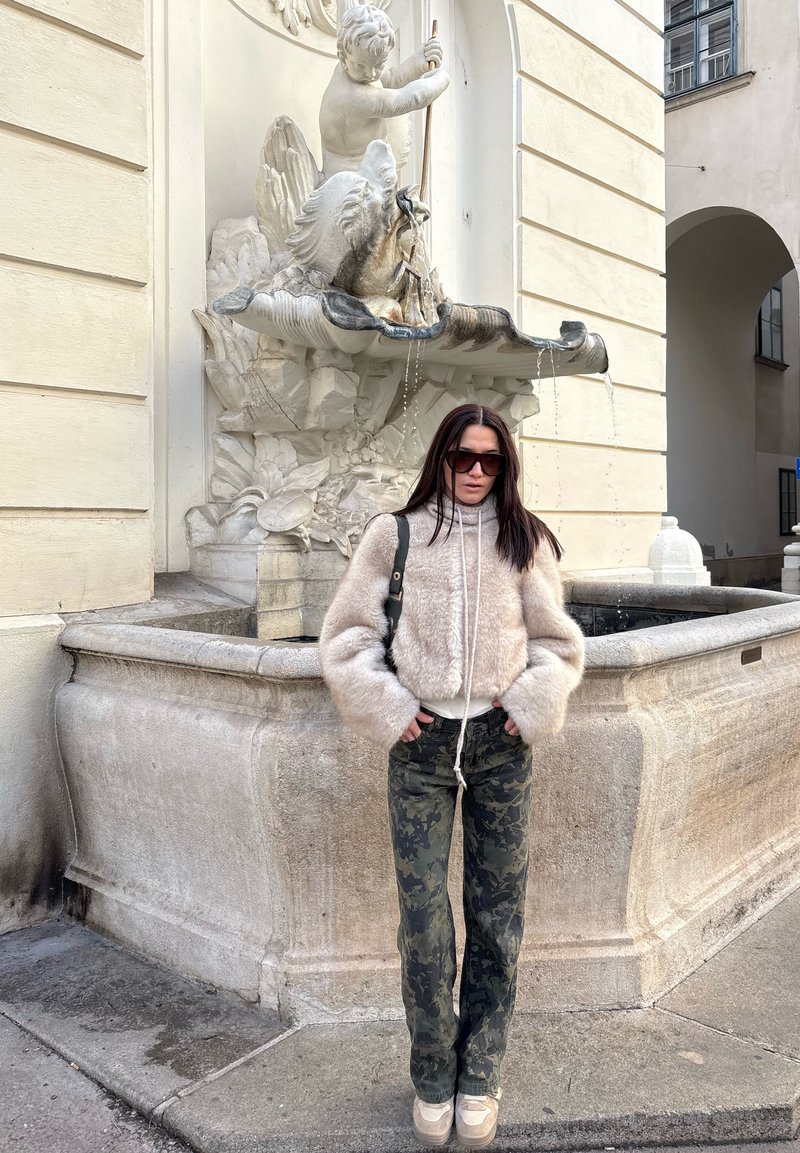 Pull crème court et flou avec des manches larges ; pantalon camouflage vert ; lunettes de soleil noires ; debout près d'une fontaine en pierre ornée d'un motif de chérubin.