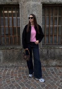 Femme en lunettes de soleil portant un haut rose, une veste noire, un jean à jambes larges et des baskets blanches, tenant un sac à main à motifs dans une rue pavée.
