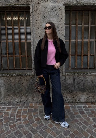 Mujer con gafas de sol, vestida con una blusa rosa, chaqueta negra, vaqueros de pierna ancha y zapatillas blancas, sosteniendo un bolso estampado en una calle empedrada.