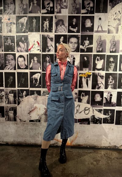 Vestido de mezclilla con bolsillos y cinturón, color azul claro, hasta la rodilla, superpuesto sobre una camisa de manga larga rosa, combinado con botas negras hasta el tobillo.