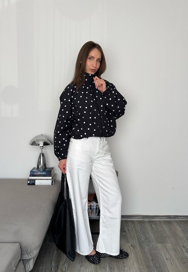 Frau trägt schwarze Jacke mit Punkten, weiße Hose und passende gepunktete Schuhe, hält eine schwarze Tasche und steht in einem minimalistischen Zimmer.