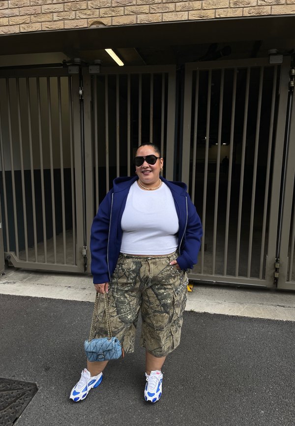 Personne portant des lunettes de soleil, une veste bleue, une chemise blanche, un short camouflage et des baskets bleu-blanc se tient à l'extérieur près de portails métalliques en tenant un sac à main bleu.