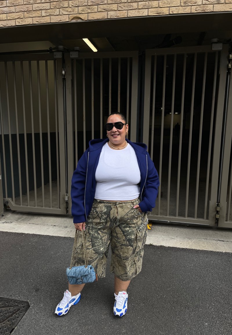 Personne portant des lunettes de soleil, une veste bleue, une chemise blanche, un short camouflage et des baskets bleu-blanc se tient à l'extérieur près de portails métalliques, tenant un sac bleu.
