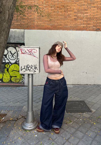 Jeans oscuros amplios con un detalle de bordado floral, combinados con una blusa rosa ajustada y una gorra gris con estampado. De pie al lado de un poste cubierto de grafitis.