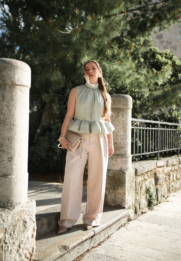 Kvinde stående udendørs nær stensøjler og metalgelænder, iført lysegrøn ærmeløs top, beige bukser med vide ben, holder en flettet clutch.