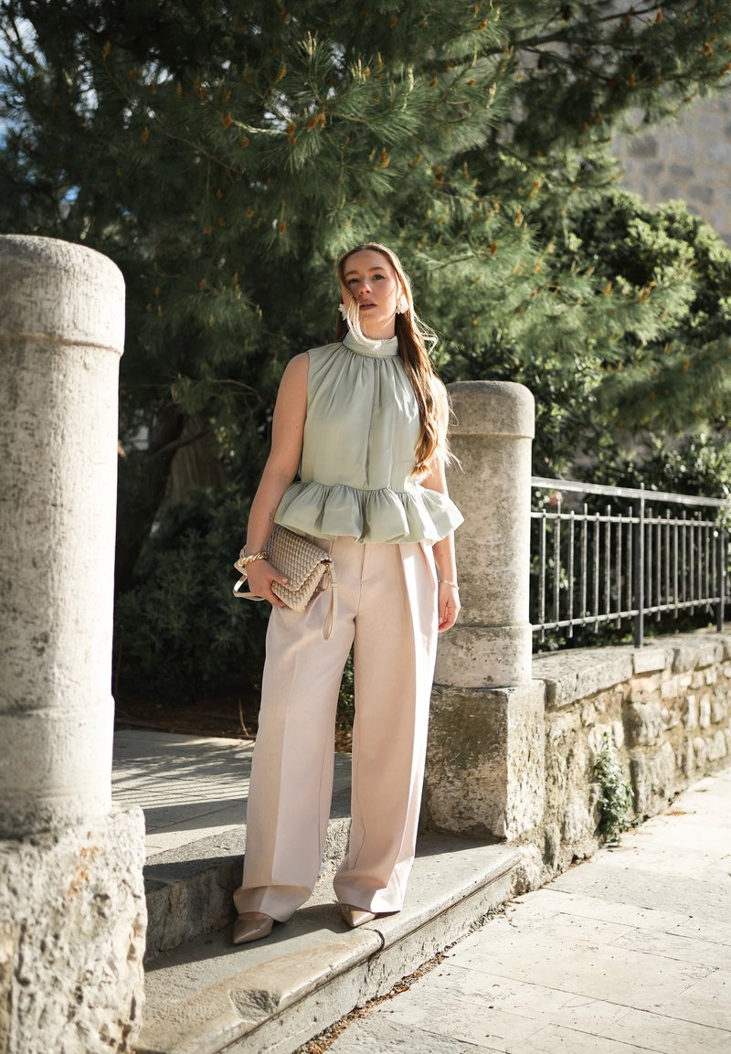 Femme debout à l'extérieur près de piliers en pierre et d'une rambarde en métal, portant un haut sans manches vert clair, un pantalon beige à jambes larges, tenant une pochette tissée.