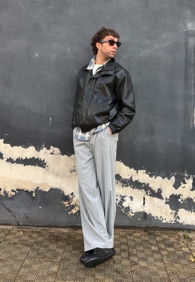 Hombre con chaqueta de cuero negra, camisa de cuadros, pantalones grises y gafas de sol apoyado contra una pared desgastada de color negro y beige.