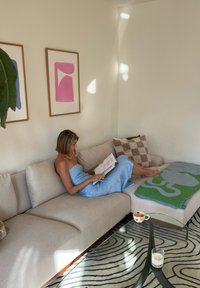 Svetlo béžová rohová sedačka s dekoračnými vankúšmi, sklenený konferenčný stolík a vzorovaný koberec. Na sedačke je prehodená tyrkysová deka.