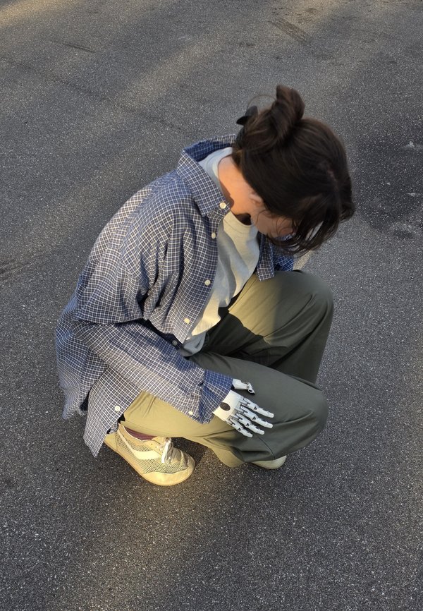 Personne accroupie sur le trottoir portant un pantalon vert et une chemise à carreaux bleue, avec une main prothétique robotique blanche visible reposant sur son genou.