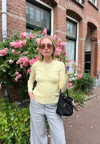 Gelber gerippter Cardigan mit Knöpfen, graue weite Stoffhosen, schwarze Bucket Bag und Sonnenbrille, posiert vor pink blühenden Büschen.