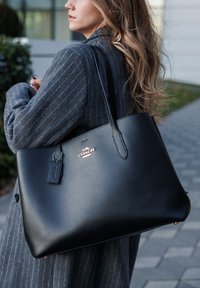 Μαύρη δερμάτινη τσάντα tote με λεία υφή, διαθέτει χρυσό λογότυπο και μια μικρή ετικέτα προσαρτημένη. Έχει σχήμα με επιμηκημένες πλευρές και ανθεκτικούς λουράκια.