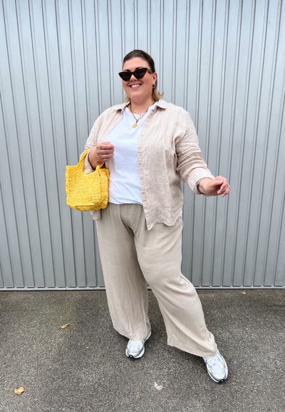 Camisa de lino beige, camiseta blanca, pantalones anchos beige, zapatillas gris, y un bolso de mimbre amarillo. Gafas de sol negras y collares de oro en capas.