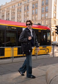 Asmuo, dėvintis pilką tekstūruotą švarką, mėlyną gobtuvą, platėjusias džinsus ir juodus sandalų, nešiojantis tamsią pašto krepšį, prie tramvajaus.