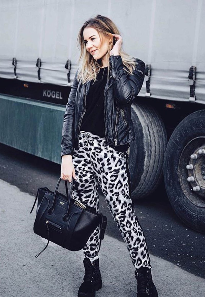 Fekete bőr kabát, fekete felső, és fehér leopárd mintás nadrág. Fekete táskát tart a markolatán; fekete harci csizmák. Városi háttér.