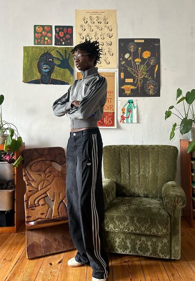 Chaqueta gris con cremallera y rayas negras, pantalones anchos negros con rayas blancas y zapatos beige. El fondo presenta una variada obra de arte y un sillón verde.