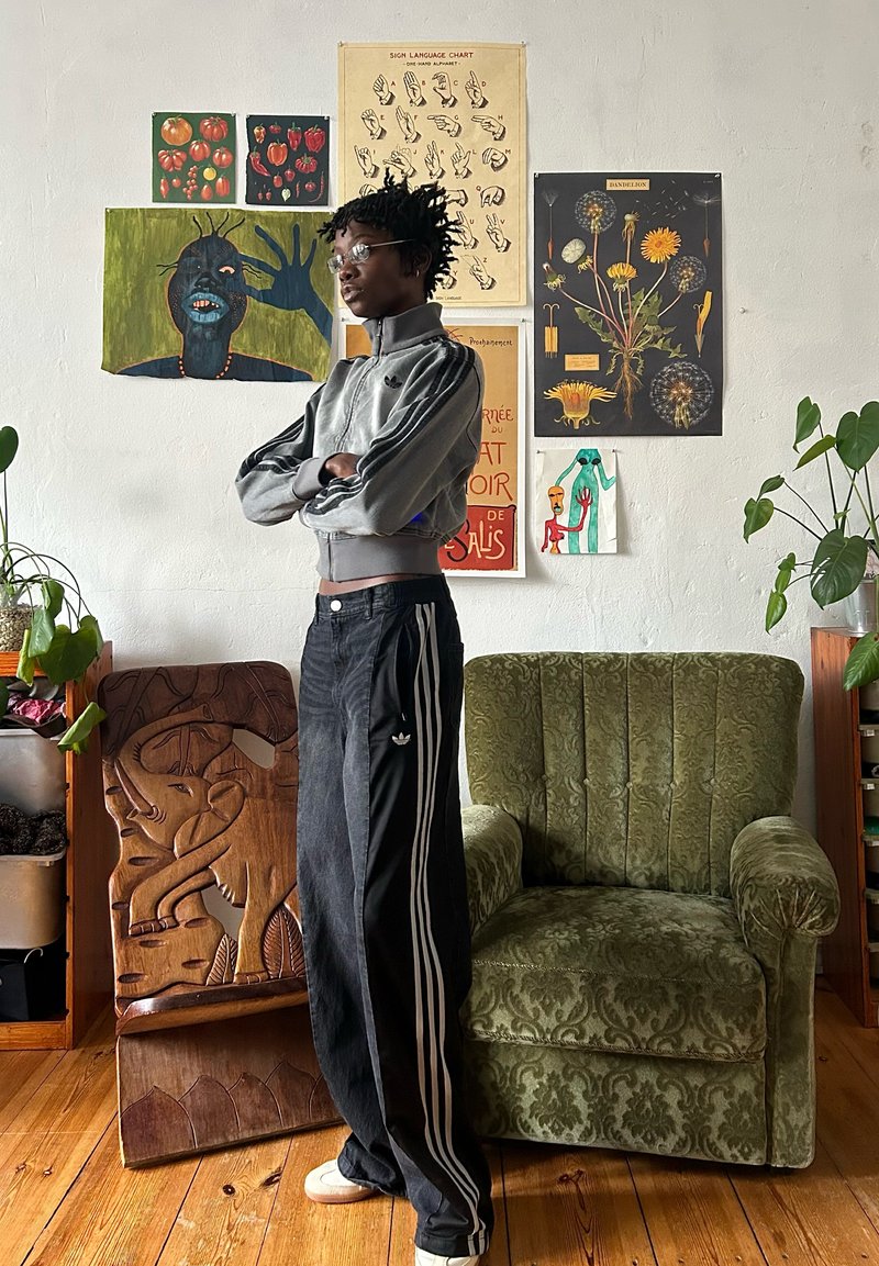 Giacca grigia con zip e strisce nere, pantaloni a gamba larga neri con strisce bianche e scarpe beige. Lo sfondo presenta opere d'arte diverse e una poltrona verde.