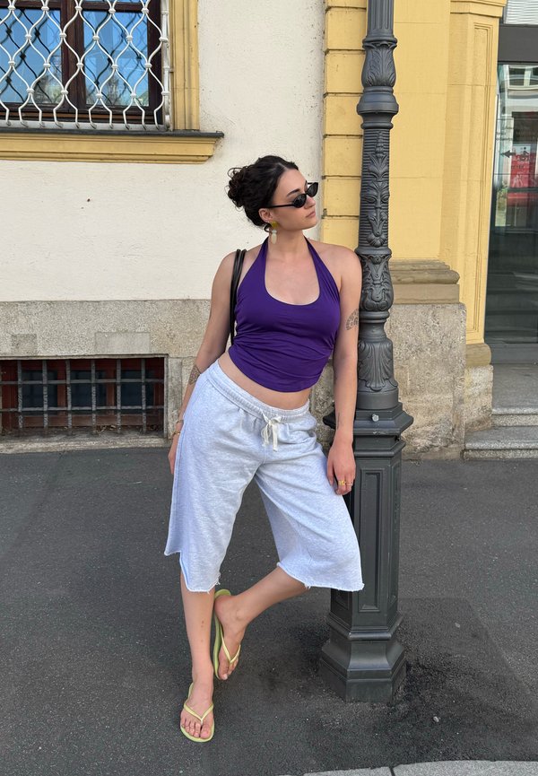 Žena v fialovom topáne s odhalenými plecami, svetlošedých krátkych nohaviciach po kolená a žltých žabkách, opierajúca sa o čierne ozdobné pouličné osvetlenie na mestskom chodníku.