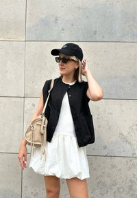 Svart knapped vest over en hvit tettsittende topp og strukturert hvit skjørt. En kremfarget veske henger fra skulderen, komplementert med solbriller og caps.