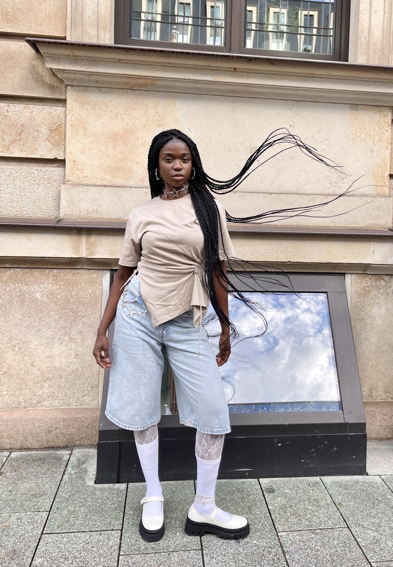 Beige Wickeloberteil, hellblaue Jeansshorts, weiße Kniestrümpfe und klobige weiße Schuhe. Lange schwarze Zöpfe wehen im Wind.