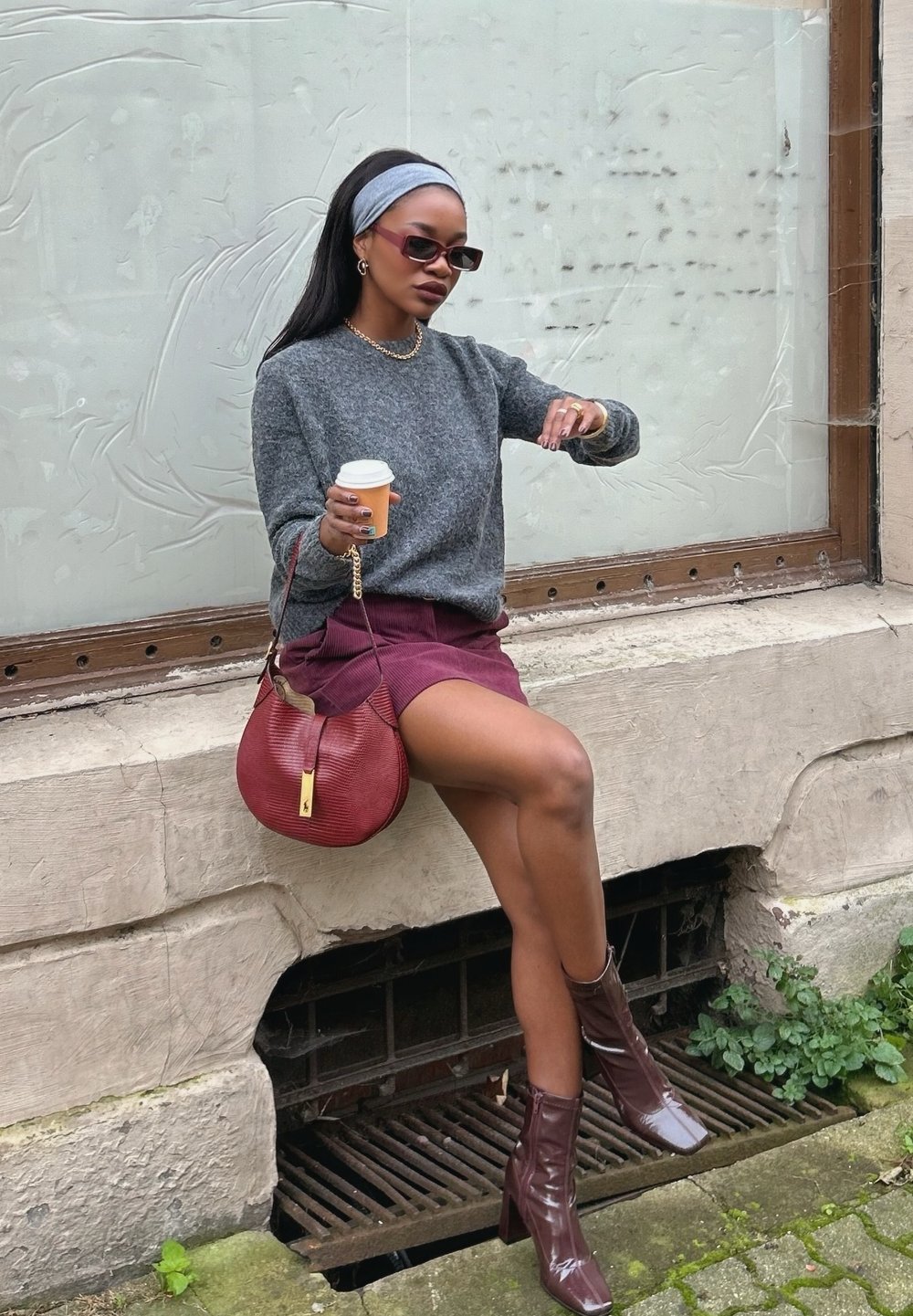 Maglione grigio, gonna bordeaux e stivaletti ankle in pelle lucida marrone. Borsa rossa testurizzata. Tiene una tazza di caffè. Seduta su un muretto di pietra.