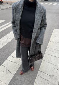 Manteau en laine gris, col roulé noir, blouse brune avec bordure en dentelle, pantalon gris évasé, sac à main brun et chaussures avec accents violets sur un passage piéton.