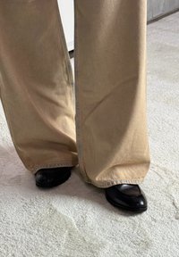 Beige, széles lábú nadrág, látható varrással, fekete, hegyes orrú cipőkkel párosítva. Könnyű szövet textúra sima felülettel.