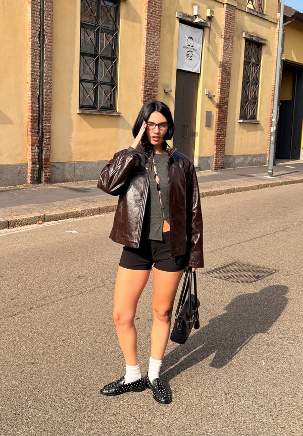 Braune glänzende Lederjacke, graues Oberteil mit Knöpfen, schwarze Shorts, weiße Socken, schwarze Schuhe mit Nieten, hält eine schwarze Tasche. Urbaner Hintergrund.