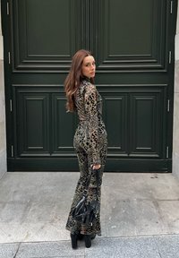 Γυναίκα με ολόσωμη φόρμα λεοπάρ και πλατφόρμες, κρατώντας μαύρη τσάντα, στέκεται σε γκρι πεζοδρόμιο μπροστά σε μεγάλη σκούρα πράσινη πόρτα.